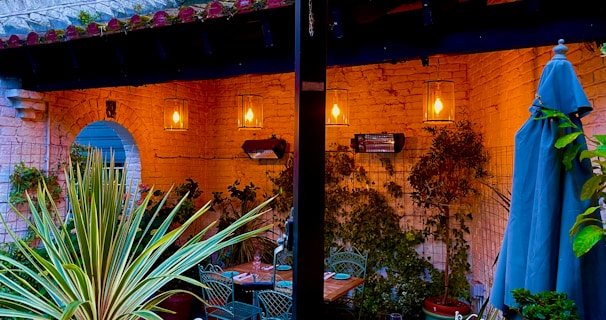 An intimate outdoor patio area adorned with warm lighting hangs against textured brick walls. The foreground features vibrant green plants and a closed patio umbrella. Wrought iron furniture is arranged around a table, creating a cozy and inviting atmosphere. Lantern-style lights cast a warm glow, enhanced by the climbing plants along the walls.