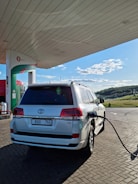 A white SUV is parked at a fuel station, connected with a fueling hose. The station has an overhead canopy and a clear sky is visible in the background. The area around the station is paved with bricks, and there is a green landscape in the distance.