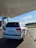 A white SUV is parked at a fuel station, connected with a fueling hose. The station has an overhead canopy and a clear sky is visible in the background. The area around the station is paved with bricks, and there is a green landscape in the distance.