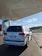A white SUV is parked at a fuel station, connected with a fueling hose. The station has an overhead canopy and a clear sky is visible in the background. The area around the station is paved with bricks, and there is a green landscape in the distance.