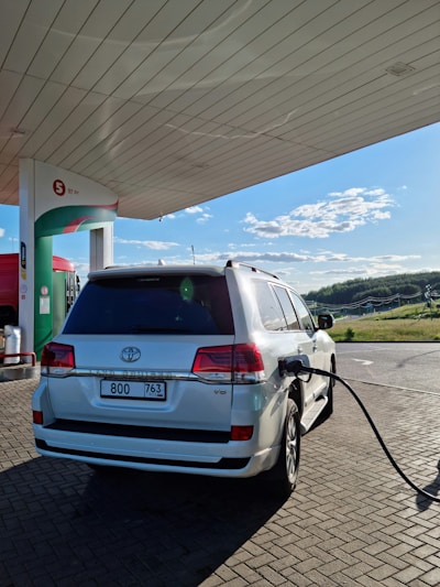 A white SUV is parked at a fuel station, connected with a fueling hose. The station has an overhead canopy and a clear sky is visible in the background. The area around the station is paved with bricks, and there is a green landscape in the distance.