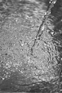 A grayscale image depicting a stream of water flowing onto a textured, rippling surface. The water appears clear, with visible bubbles and a smooth, continuous motion.