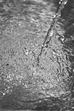 A grayscale image depicting a stream of water flowing onto a textured, rippling surface. The water appears clear, with visible bubbles and a smooth, continuous motion.