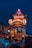 A vibrant festival procession features an ornately decorated float with lanterns, a figure in traditional red clothing, and musicians playing drums. The scene is set against a backdrop of traditional architecture, with spectators gathered around, many wearing masks.