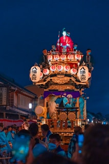 A vibrant festival procession features an ornately decorated float with lanterns, a figure in traditional red clothing, and musicians playing drums. The scene is set against a backdrop of traditional architecture, with spectators gathered around, many wearing masks.