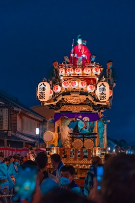 A vibrant festival procession features an ornately decorated float with lanterns, a figure in traditional red clothing, and musicians playing drums. The scene is set against a backdrop of traditional architecture, with spectators gathered around, many wearing masks.