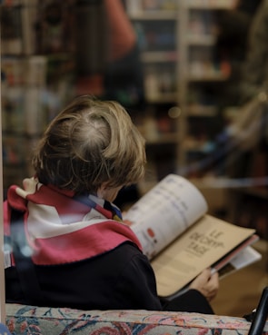 A person, likely a child, is seen from behind sitting on a couch with a colorful fabric pattern. They are reading a book in a cozy indoor setting, possibly a bookstore or library. The individual's hair is light brown and somewhat tousled, and they are wearing a jacket with a red scarf or accessory. The focus is on the book they are reading, which has text and images on the open pages.