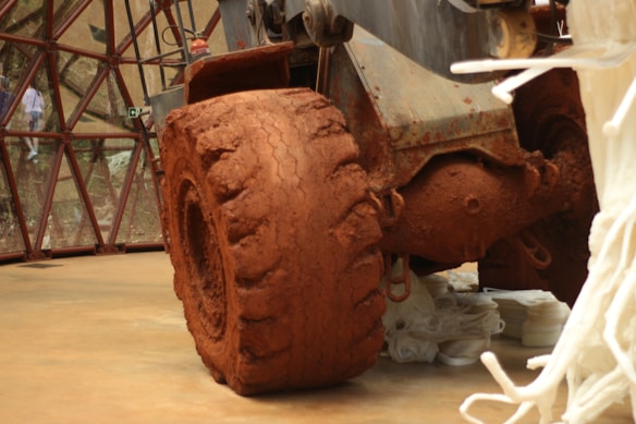 A large, heavily dirt-covered tire attached to industrial machinery dominates the foreground. The machinery is placed indoors on a smooth, light brown floor. In the background, there is a glass-paneled wall with a geometric framework, outside of which some greenery can be seen. White abstract sculptures or installations surround the machinery, adding an artistic touch.
