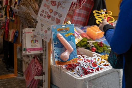 A market stall displaying a diverse assortment of novelty goods. Prominent among the items is a package labeled 'Water Gun' shaped humorously. Surrounding the main item are various party decorations, including heart and birthday-themed designs, as well as decorative items like glitter glasses with the number 2022. A person in a blue top is seen browsing the items, holding a sparkly butterfly wand.