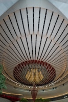 An architectural interior with a striking, circular ceiling design features alternating dark and light panels. A large, hanging decorative structure is in the center, resembling an abstract chandelier. The area is well-lit with ambient lighting, and there are touches of green from small trees or plants.
