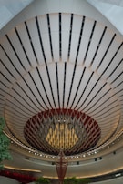 An architectural interior with a striking, circular ceiling design features alternating dark and light panels. A large, hanging decorative structure is in the center, resembling an abstract chandelier. The area is well-lit with ambient lighting, and there are touches of green from small trees or plants.