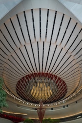 An architectural interior with a striking, circular ceiling design features alternating dark and light panels. A large, hanging decorative structure is in the center, resembling an abstract chandelier. The area is well-lit with ambient lighting, and there are touches of green from small trees or plants.