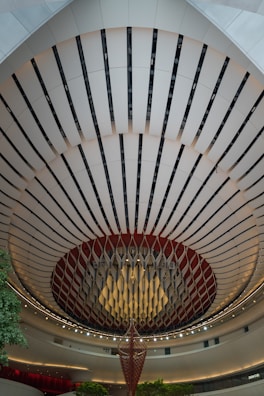 An architectural interior with a striking, circular ceiling design features alternating dark and light panels. A large, hanging decorative structure is in the center, resembling an abstract chandelier. The area is well-lit with ambient lighting, and there are touches of green from small trees or plants.