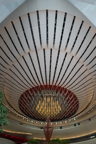 An architectural interior with a striking, circular ceiling design features alternating dark and light panels. A large, hanging decorative structure is in the center, resembling an abstract chandelier. The area is well-lit with ambient lighting, and there are touches of green from small trees or plants.