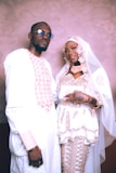 A couple is dressed in traditional wedding attire, with the man wearing a white outfit with pink geometric patterns and sunglasses, and the woman wearing a white gown with intricate lace and embroidery, along with a headscarf and veil. She has intricate henna designs on her hands and is holding a card.