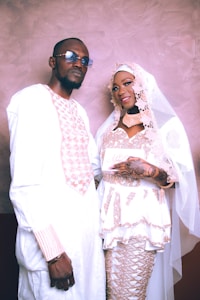 A couple is dressed in traditional wedding attire, with the man wearing a white outfit with pink geometric patterns and sunglasses, and the woman wearing a white gown with intricate lace and embroidery, along with a headscarf and veil. She has intricate henna designs on her hands and is holding a card.