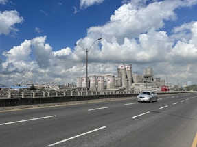 A large industrial facility with numerous cylindrical storage tanks and chimneys is seen beside a wide, multi-lane highway. One car is on the road, while the sky above is bright and filled with fluffy clouds.