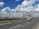 A large industrial facility with numerous cylindrical storage tanks and chimneys is seen beside a wide, multi-lane highway. One car is on the road, while the sky above is bright and filled with fluffy clouds.
