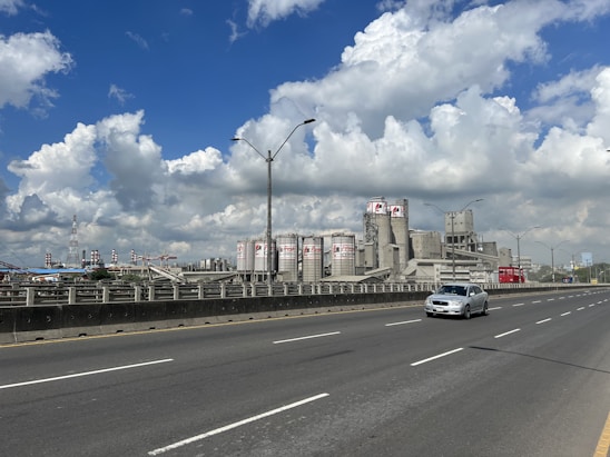 A large industrial facility with numerous cylindrical storage tanks and chimneys is seen beside a wide, multi-lane highway. One car is on the road, while the sky above is bright and filled with fluffy clouds.