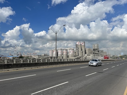 A large industrial facility with numerous cylindrical storage tanks and chimneys is seen beside a wide, multi-lane highway. One car is on the road, while the sky above is bright and filled with fluffy clouds.