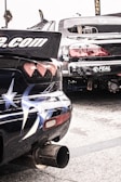 Two modified, sports cars are parked in a lot, showcasing stickers and elaborate paint designs, including stars on one car's rear. The rear ends of the vehicles are prominent, emphasizing their aerodynamic modifications. Their exhaust systems are visible, suggesting potential for high performance.