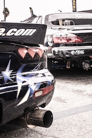 Two modified, sports cars are parked in a lot, showcasing stickers and elaborate paint designs, including stars on one car's rear. The rear ends of the vehicles are prominent, emphasizing their aerodynamic modifications. Their exhaust systems are visible, suggesting potential for high performance.