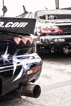 Two modified, sports cars are parked in a lot, showcasing stickers and elaborate paint designs, including stars on one car's rear. The rear ends of the vehicles are prominent, emphasizing their aerodynamic modifications. Their exhaust systems are visible, suggesting potential for high performance.