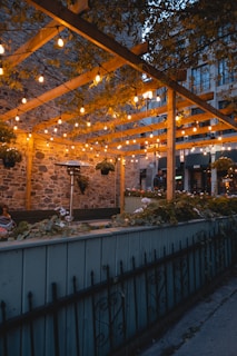 Голяма дървена конструкция, осветена през нощта, с хора, които вечерят под нея в София.