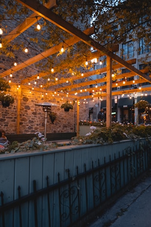 Голяма дървена конструкция, осветена през нощта, с хора, които вечерят под нея в София.