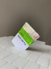 A rectangular block covered with silver and green foil is placed on a tiled surface. The block has a label with the text 'CALL KEN LIM T.S.' printed on a white background. The tiles are glossy and white, arranged in a slightly offset manner, while the wall in the background is plain and grey.