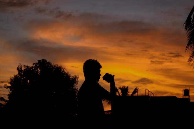 A silhouette of a person holding a camera stands against a vivid sunset sky. The horizon features dark trees and some structures, creating a striking contrast with the bright orange and yellow hues of the sunset. Wispy clouds are scattered across the sky, adding texture to the scene.