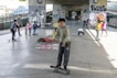 Children are skateboarding and playing under a bridge in an urban environment. A young boy in the foreground is smiling while standing on a skateboard. Several other children and an adult are visible, some actively skating and others watching. Graffiti decorates the pillars and walls, and there is a lively atmosphere.