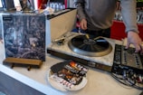 A person is operating a turntable, which is set on a counter along with a mixer and some vinyl records. An album cover stands upright nearby, showing artistic black and white imagery. Several promotional flyers or cards are scattered on the counter.