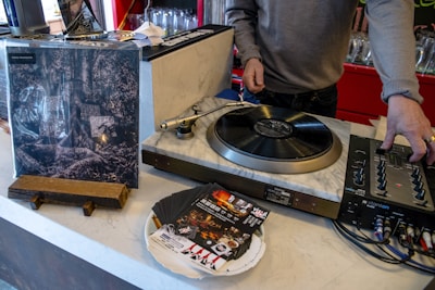 A person is operating a turntable, which is set on a counter along with a mixer and some vinyl records. An album cover stands upright nearby, showing artistic black and white imagery. Several promotional flyers or cards are scattered on the counter.