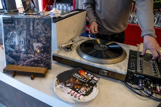 A person is operating a turntable, which is set on a counter along with a mixer and some vinyl records. An album cover stands upright nearby, showing artistic black and white imagery. Several promotional flyers or cards are scattered on the counter.