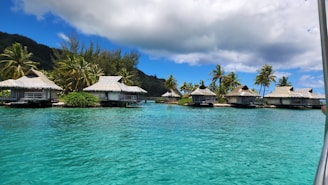 A serene tropical scene featuring several overwater bungalows with thatched roofs situated along a turquoise lagoon. Lush green palm trees and dense foliage provide a picturesque backdrop, while fluffy white clouds drift across a bright blue sky.