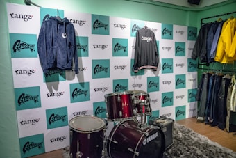 A retail display features a drum set and a clothing lineup against a wall covered in alternating logos from 'range' and another brand. On the wall, a blue hoodie and a black jacket are showcased. A clothing rack to the right holds various jackets and jeans in colors like yellow, blue, and black. The flooring is a mixed-texture with wood and a gray plush carpet.