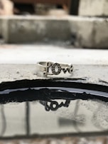 A silver ring featuring the word 'love' resting on a rough concrete surface. The ring's reflection is visible in a small puddle of water beneath it, adding depth to the composition. The background is blurred, enhancing the focus on the ring.