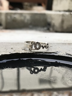 A silver ring featuring the word 'love' resting on a rough concrete surface. The ring's reflection is visible in a small puddle of water beneath it, adding depth to the composition. The background is blurred, enhancing the focus on the ring.