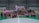 A group of people are gathered on an indoor futsal court. Some are holding prizes and trophies, indicating that they are part of a futsal tournament or competition. There is a large banner in the background with text related to the event, featuring a person's image.