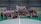 A group of people are gathered on an indoor futsal court. Some are holding prizes and trophies, indicating that they are part of a futsal tournament or competition. There is a large banner in the background with text related to the event, featuring a person's image.