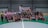 A group of people are gathered on an indoor futsal court. Some are holding prizes and trophies, indicating that they are part of a futsal tournament or competition. There is a large banner in the background with text related to the event, featuring a person's image.