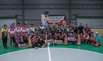 A group of people are gathered on an indoor futsal court. Some are holding prizes and trophies, indicating that they are part of a futsal tournament or competition. There is a large banner in the background with text related to the event, featuring a person's image.