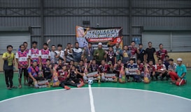 A group of people are gathered on an indoor futsal court. Some are holding prizes and trophies, indicating that they are part of a futsal tournament or competition. There is a large banner in the background with text related to the event, featuring a person's image.