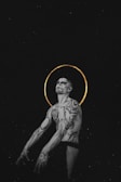 A tattooed person is depicted with intricate and geometric designs covering the upper body. A golden halo encircles the head, providing a stark contrast against the dark background, adding an ethereal quality to the image.