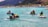 Four people riding jet skis on clear turquoise water with lush green mountains and bungalows in the background.