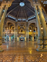 An ornate, expansive hall with intricately designed columns featuring detailed carvings and patterns. The ceiling is domed with a skylight at the center, surrounded by elaborate architectural designs. A large chandelier hangs from the ceiling, and the floor displays elaborate tile work. In the background, groups of people are gathering, indicating the space is accessible to the public.