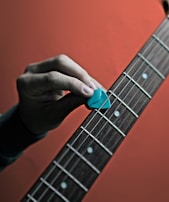 A hand holding a blue guitar pick is positioned above the strings of a guitar. The fretboard features visible white dots as position markers, and the background is a gradient of orange hues.