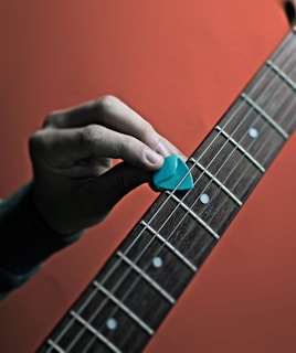 A hand holding a blue guitar pick is positioned above the strings of a guitar. The fretboard features visible white dots as position markers, and the background is a gradient of orange hues.
