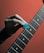 A hand holding a blue guitar pick is positioned above the strings of a guitar. The fretboard features visible white dots as position markers, and the background is a gradient of orange hues.